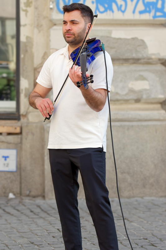 26 - Young person playing a bright blue electric violin on a cobblestone street